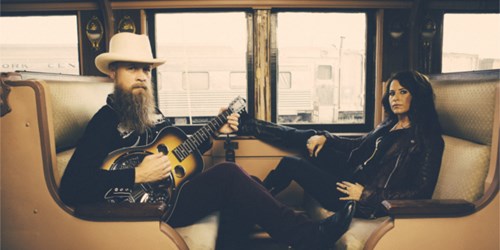 The Grahams - Man and woman sitting opposite each other on a train, Man is holding a guitar.